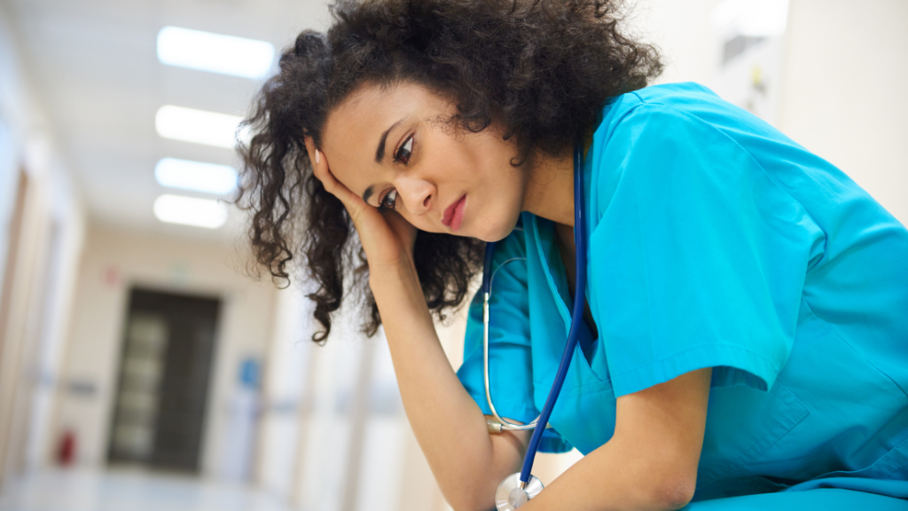 A nurse wearing scrubs and a stethoscope sits in a hospital hallway, holding her forehead with one hand.